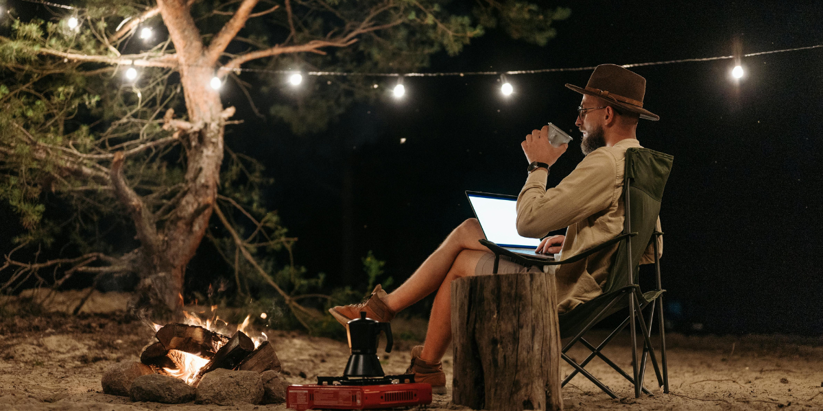 Person typing on a laptop near a cozy campfire surrounded by tall trees